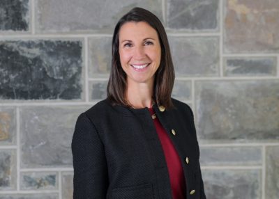 A person with shoulder-length dark hair stands smiling before a wall of grey stone.