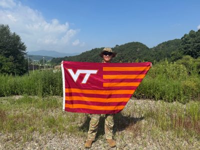 A serviceman in a camouflage uniform and sunglasses stands in front of a mountain view holding a Virginia Tech flag while smiling.