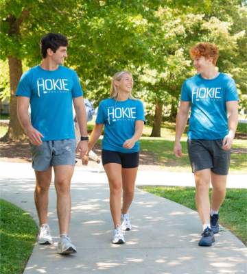 Three smiling people walk in the dappled light of trees while wearing shorts and blue T-shirts that read "I am the Hokie Movement" in white letters.