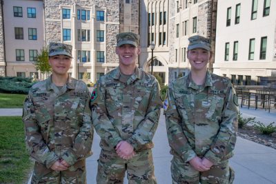 Cadets in uniform stand smiling in front of a Hokie Stone building. 
