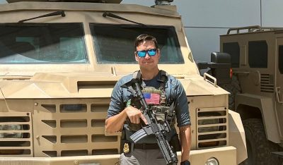 A man with a badge holding a rifle stands in front of a military vehicle.
