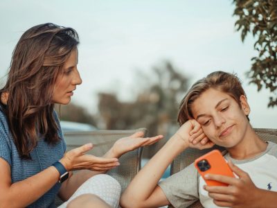 A teenager smirks while staring at a bright-orange phone. To his left his mother looks  at him, gesturing.