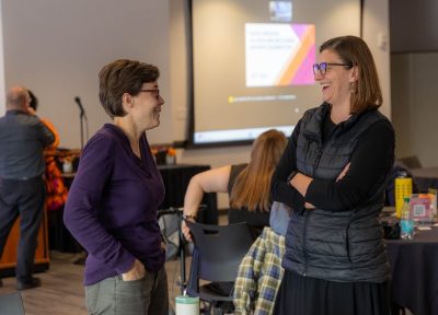 Two individuals stand smiling, talking to each other in a conference room.